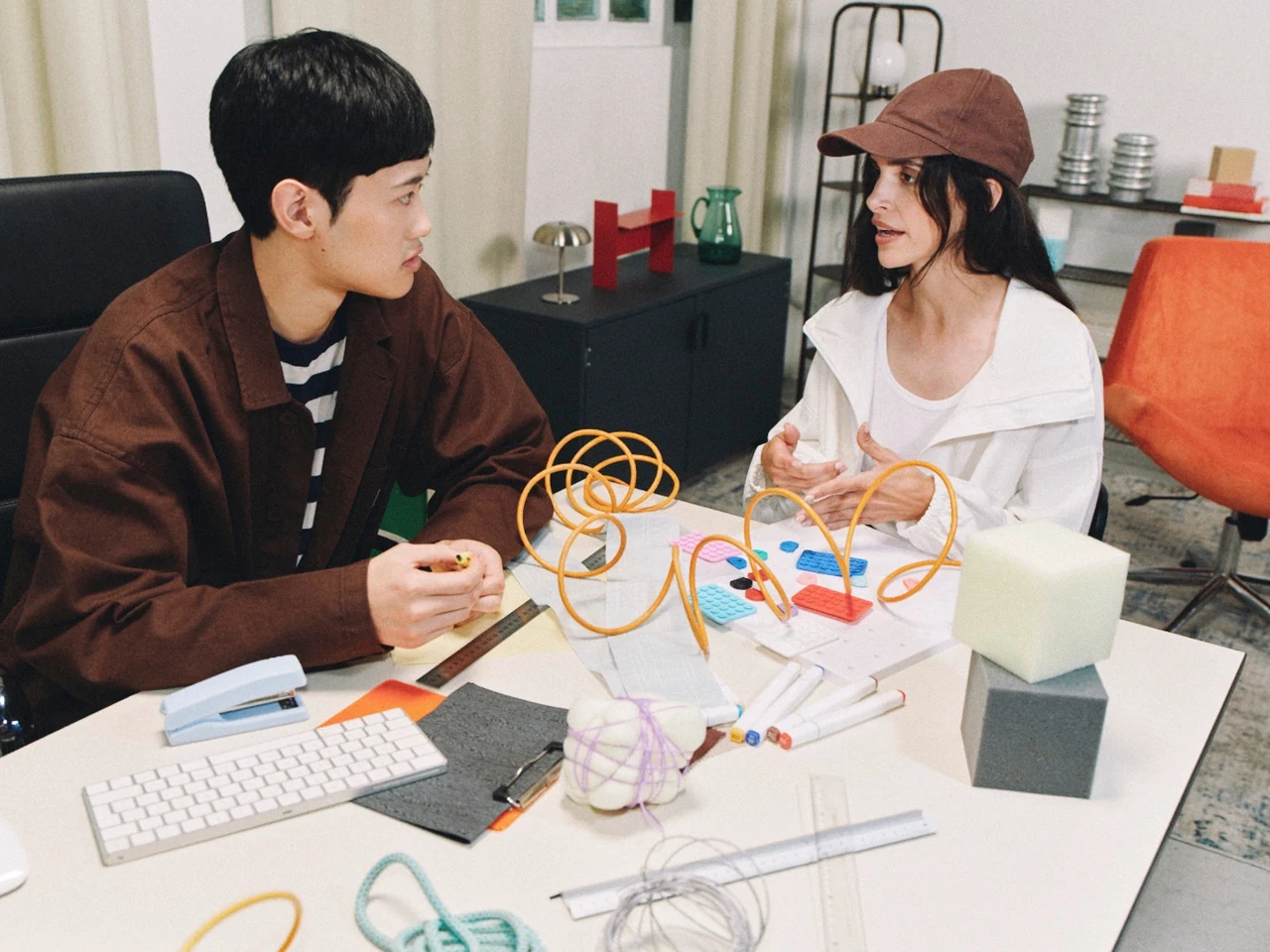 Two people talking across a desk covered with creative materials.